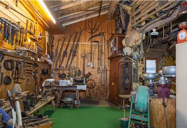 A room full of old tools and agricultural equipment. The walls are decorated with various utensils and a wooden shelf.