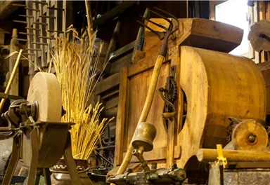 A historic craft tool with wooden details stands in an old shed. Next to the tool, there are dried stalks and various tools visible.