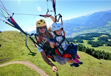A man and a child glide in paragliding over a green landscape with mountains in the background. The sky is clear and blue, and the weather is sunny.