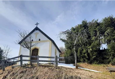 A small, white chapel stands on a hill, surrounded by trees. In the background, the blue sky and a gentle landscape can be seen.