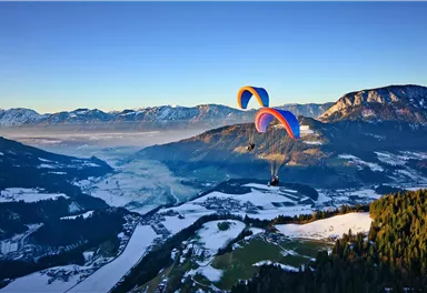 Two paragliders glide over a charming alpine landscape. In the background, snow-covered mountains and a clear sky can be seen.