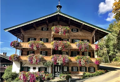 Ein schönes Holzhaus mit zahlreichen bunten Blumen in den Fenstern. Der Himmel ist klar und blau, umgeben von grüner Natur.
