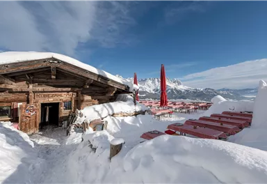 Eine gemütliche Berghütte im Schnee mit roten Tischen im Freien. Im Hintergrund sind schneebedeckte Berge und ein blauer Himmel zu sehen.
