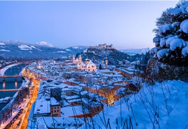 A picturesque town in winter, covered with snow and surrounded by mountains. The lights of the town reflect in the calm water of the river.