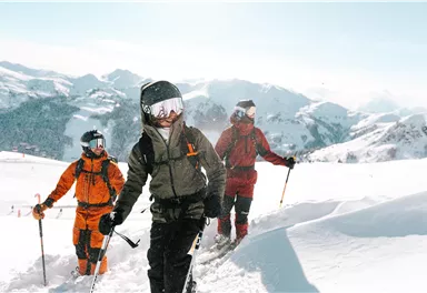 Three people are hiking through a snow-covered mountain landscape. The sun is shining, and the surroundings are picturesque with tall mountains in the background.