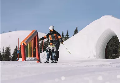 A ski instructor is helping a child ski on a snow-covered slope. The scene shows sunny weather with a snowy landscape in the background.
