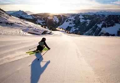 A skier is skiing down a wide, sunny slope. In the background, snow-covered mountains and a clear sky can be seen.