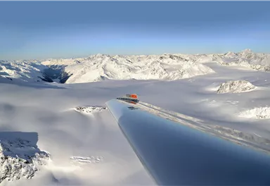 Eine beeindruckende Schneelandschaft mit majestätischen Bergen im Hintergrund. Im Vordergrund ist ein schimmernder, reflektierender Flügel eines Flugzeugs zu sehen.
