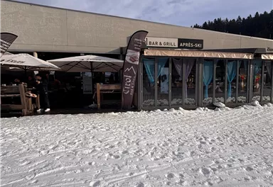 Ein après-ski Restaurant mit einer Terrasse in der Schnee. Es gibt Sonnenschirme und die Umgebung ist winterlich und bergig.