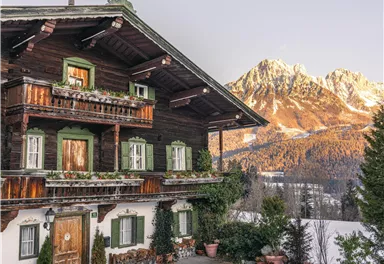 Ein traditionelles Holzhaus im Schnee mit Balkonen und Blumen. Im Hintergrund sind schneebedeckte Berge zu sehen.