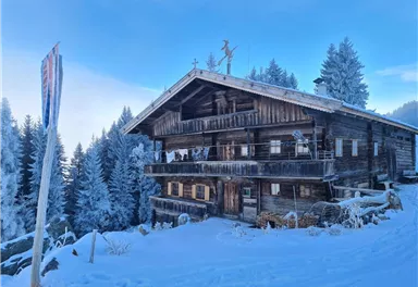 A traditional wooden house in the snow, surrounded by tall, frosty fir trees. The sky is blue and radiates clear winter air.