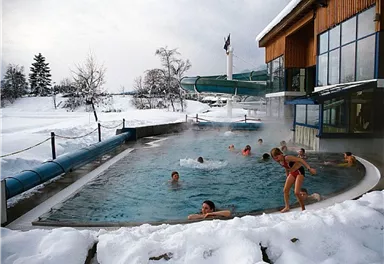 An outdoor pool in a winter setting, surrounded by snow. Several people are enjoying the warm water and there is a slide next to the pool.