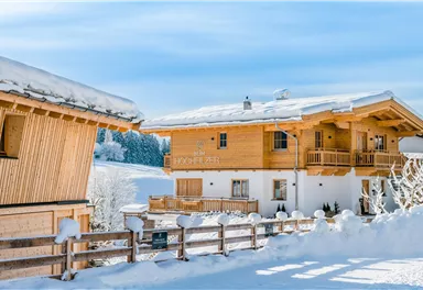 Two cozy wooden chalets in the snow, surrounded by a wintry landscape. The sky is clear and blue, creating a peaceful atmosphere.