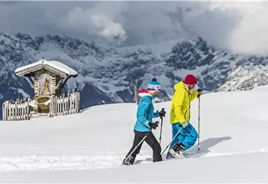 Two people are snowshoeing through the snowy landscape. In the background, impressive mountains and a small cabin can be seen.