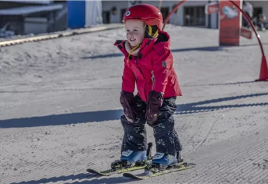 Ein fröhliches Kind in einem roten Schneeanzug fährt auf Skiern. Es steht auf einer verschneiten Piste mit einem blauen Himmel im Hintergrund.