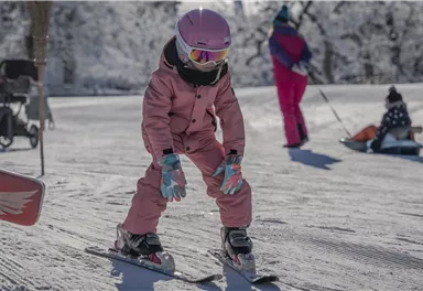 Ein kleines Mädchen in einer rosa Skijacke lernt das Skifahren auf einer verschneiten Piste. Im Hintergrund sind weitere Skifahrer und eine verschneite Landschaft zu sehen.