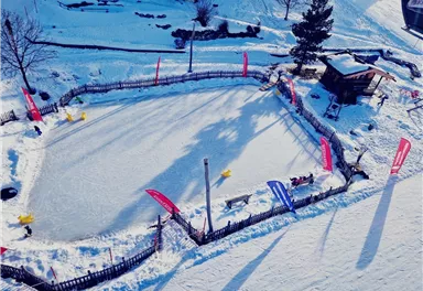 Eine Eislaufbahn umgeben von Schnee und Bäumen. Bunte Fahnen wehen in der kalten Winterluft.