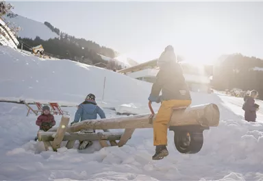 Kinder spielen im Schnee auf einem hölzernen Spielgerät. Die Sonne scheint und die Berge sind im Hintergrund sichtbar.