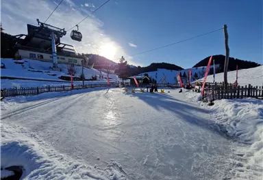 Eine Eisbahn im Schnee mit Blick auf Berge und einen Skilift. Der Himmel ist klar und die Sonne scheint.