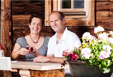 A couple sits at a wooden table in front of a rustic cabin. They are smiling and holding a glass in their hands, surrounded by colorful flowers.