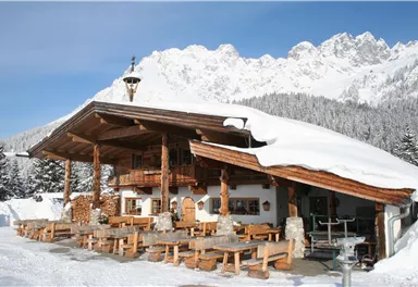 A cozy mountain cabin in the snow with a wooden terrace and tables. In the background, snow-capped mountains can be seen.