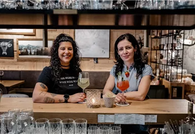 Two friendly female bartenders stand at a wooden bar holding colorful cocktails in their hands. In the background, glasses and decorative elements can be seen.