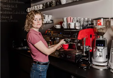 A barista is standing at a coffee machine preparing a coffee. In the background, cups and other kitchen utensils can be seen.
