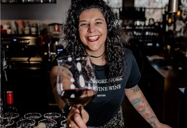 A friendly bartender smiles behind a counter and holds a wine glass. In the background, glasses and a cozy bar can be seen.