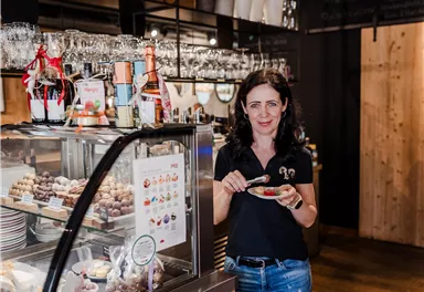 A woman stands in a café presenting a dessert platter. In the background, many delicious sweets and drinks can be seen.