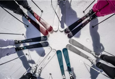 A group of skiers stands in the snow, forming a circle with their skis. The skis are in different colors and show the marks of skiing on the snow.
