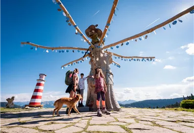 A group of people stands under a large tree as a work of art. In the background, a bright red lighthouse and a beautiful mountain landscape can be seen.