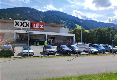 A furniture store with the inscription "XXX Lutz" on the facade. In front of the building, several cars are parked in a large parking lot, with a green landscape in the background.
