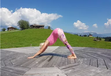 Eine Frau macht Yoga auf einer Holzplattform in der Natur. Der Himmel ist blau mit weißen Wolken und im Hintergrund sind Hügel zu sehen.