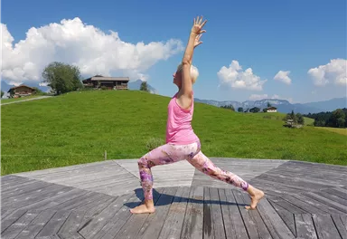 Eine Frau in Yogahaltung steht auf einer Holzplattform im Freien. Im Hintergrund sind grüne Hügel und blauer Himmel mit weißen Wolken zu sehen.