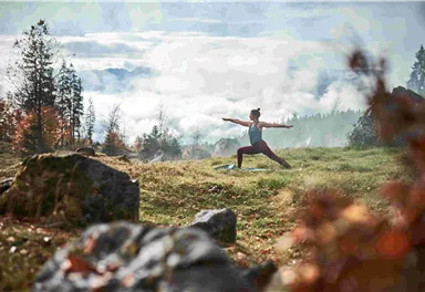 A person is doing yoga in a picturesque, wooded landscape. Gentle hills and mist can be seen in the background.