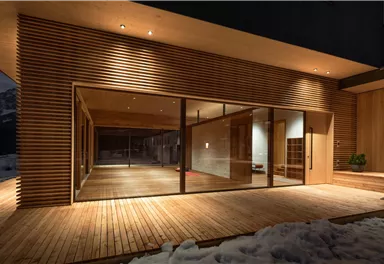 Ein modernes Haus mit großen Glasfenstern und Holzverkleidung. Der Außenbereich hat einen Holzfußboden und ist teilweise schneebedeckt.