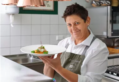 A chef in a modern kitchen proudly holds an elegant plate with a dish. The atmosphere is friendly and welcoming.