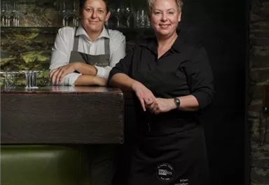 Two people are standing at a bar in a cozy restaurant. In the background, shelves with drinks are visible.