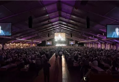 A large crowd in a tent during an event. Contributions from a speaker are displayed on screens.