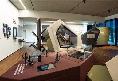 Ein modernes Ausstellungszimmer mit interaktiven Displays und Ausstellungsstücken. Es gibt mehrere Monitoren, Kopfhörer und geometrische Strukturen auf dem Tisch.