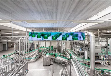 Eine moderne Produktionsanlage mit vielen Rohren und Förderbändern. Im Hintergrund ist ein großer Bildschirm zu sehen, der Informationen anzeigt.