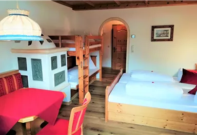 Ein gemütliches Zimmer mit einem Etagenbett und zwei Einzelbetten. Ein runder Tisch mit roten Stühlen und ein traditioneller Ofen verleihen dem Raum eine warme Atmosphäre.