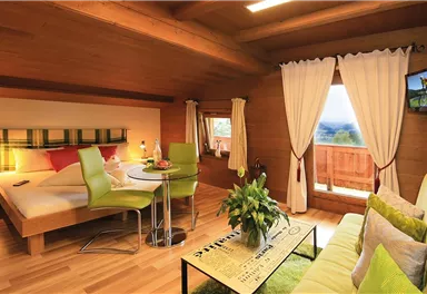 A cozy room with wooden walls, a double bed, and a small dining area. Large windows offer a view of nature.