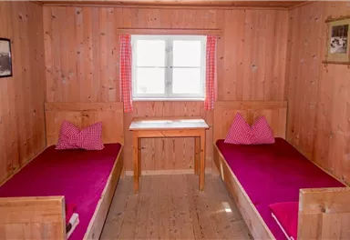 Ein gemütliches Schlafzimmer mit zwei Einzelbetten und einem Tisch in der Mitte. Die Wände sind aus Holz und es gibt Fenster mit karierten Vorhängen.