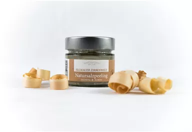 A glass of natural salt scrub stands against a white background. Next to it lie some wood shavings.