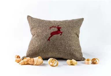 An elegiac pillow made of brown fabric with a red, embroidered reindeer. Wooden cubes and shavings lie around the pillow.