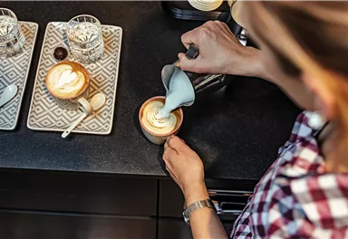 A person is pouring milk into a cup of coffee using a milk frother. Several cups and glasses are on the table.