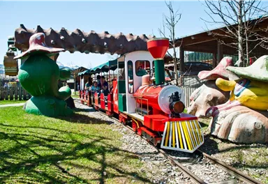A colorful train travels through an amusement park. In the background, large, creative sculptures can be seen.