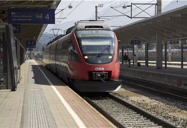 Ein roter Zug der ÖBB steht am Bahnhof. Die Gleise sind sichtbar und die Architektur des Bahnhofs ist modern.