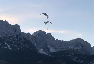 Two paragliders fly over majestic mountains. The sky is clear and showcases the beauty of nature.